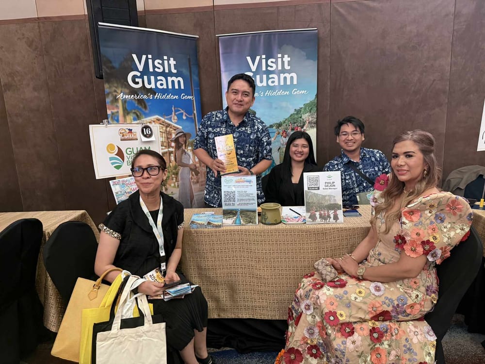 The Guam Visitors Bureau booth at seller table 16, with two Visit Guam roll-up banners and staff in Guam-print shirts meeting buyers.